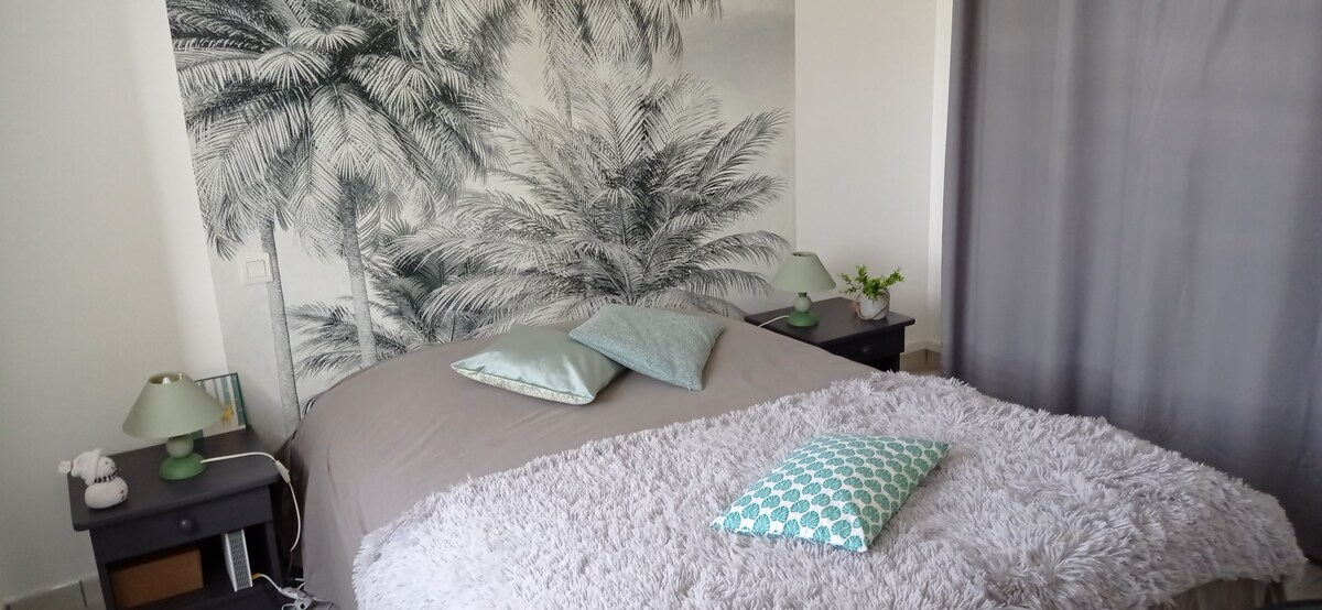 A spacious bed is positioned against a mural featuring palm trees, creating a calming backdrop. Soft pillows are arranged on the bed, with a textured throw blanket adding comfort. Two bedside lamps provide gentle illumination beside dark wooden nightstands, while a small green plant adds a touch of nature.