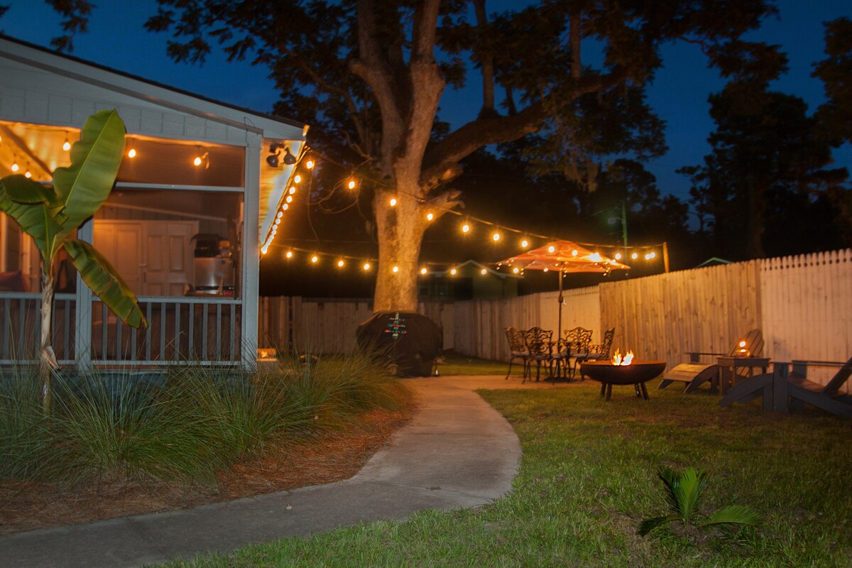 The outdoor area is illuminated by string lights, casting a warm glow over the space. A winding path leads to a fire pit surrounded by seating. A patio umbrella provides shade over a dining area, with a grill nearby and a large tree shading the scene.