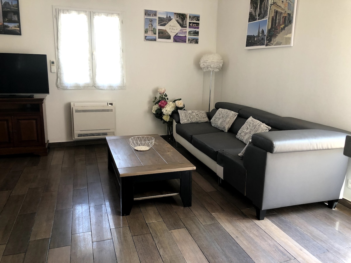 A welcoming living area features a grey sectional sofa, a wooden coffee table, and a television. Natural light enters through two windows adorned with sheer curtains. Decorative flowers are placed next to a standing lamp, and wall art enhances the space.