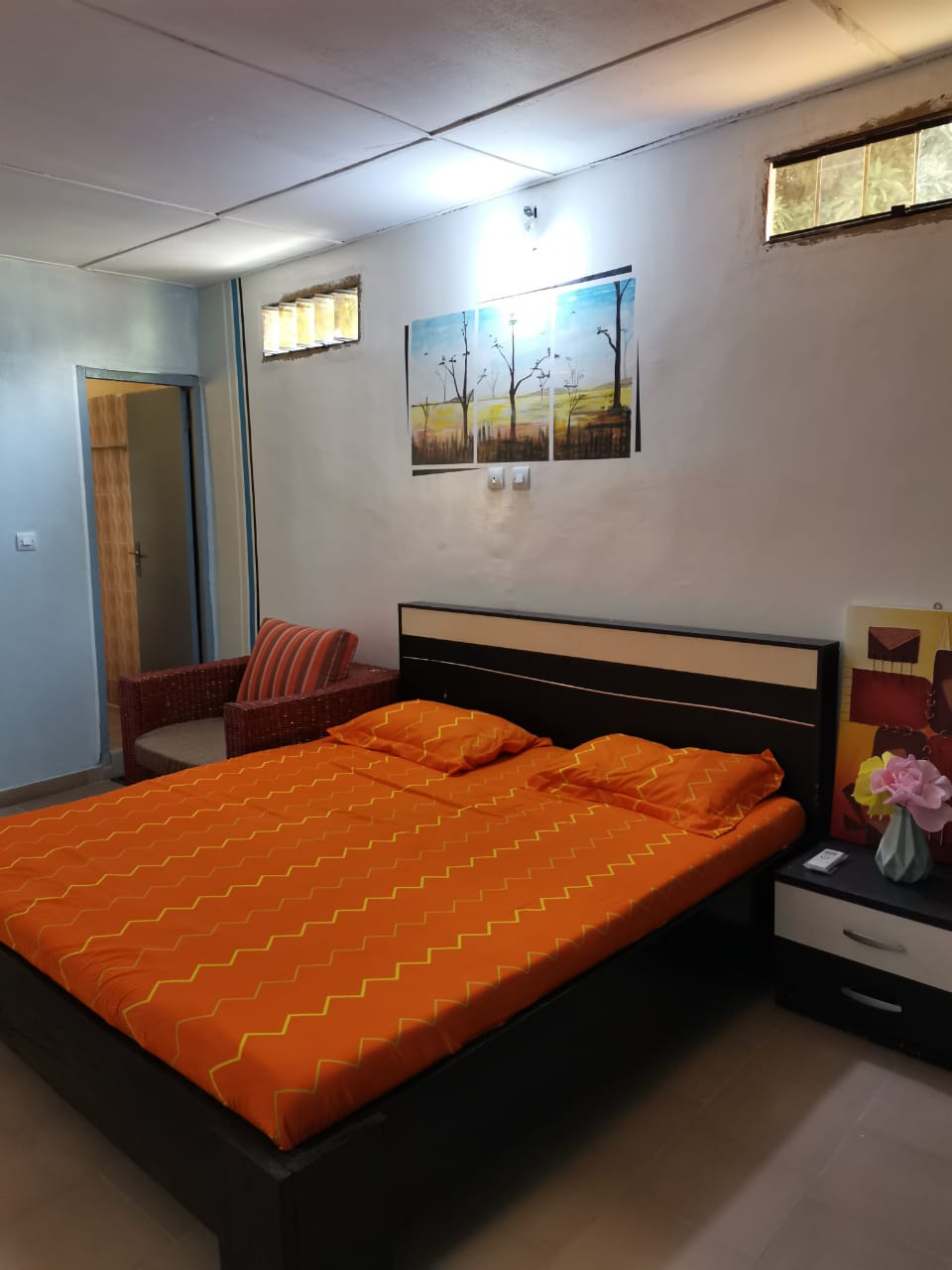 A cozy sleeping area features a spacious bed covered with an orange patterned blanket. A small nightstand sits beside the bed. A chair with a red and white striped cushion is positioned against the wall. Natural light filters through two windows near the ceiling.