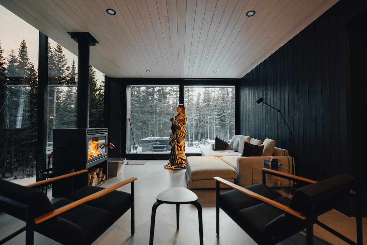 A spacious living area features large panoramic windows overlooking a forest. A sectional sofa is positioned near a modern wood fireplace. Two black armchairs are located in the foreground, enhancing the tranquil indoor environment.