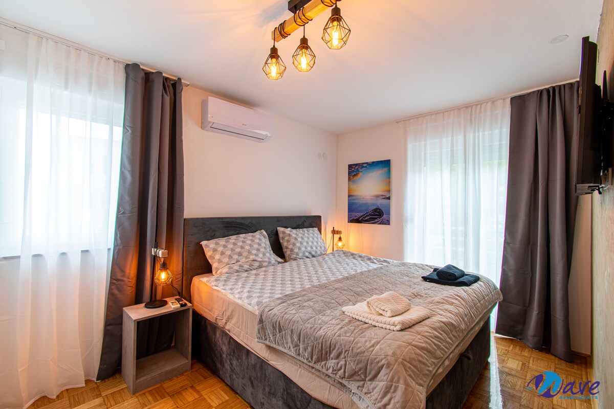 A modern bedroom features a spacious bed with a quilted comforter and accent pillows. Natural light filters through sheer curtains, while dark drapes contrast the light walls. A small bedside table holds a lamp, and a flat-screen TV is mounted on the opposite wall.