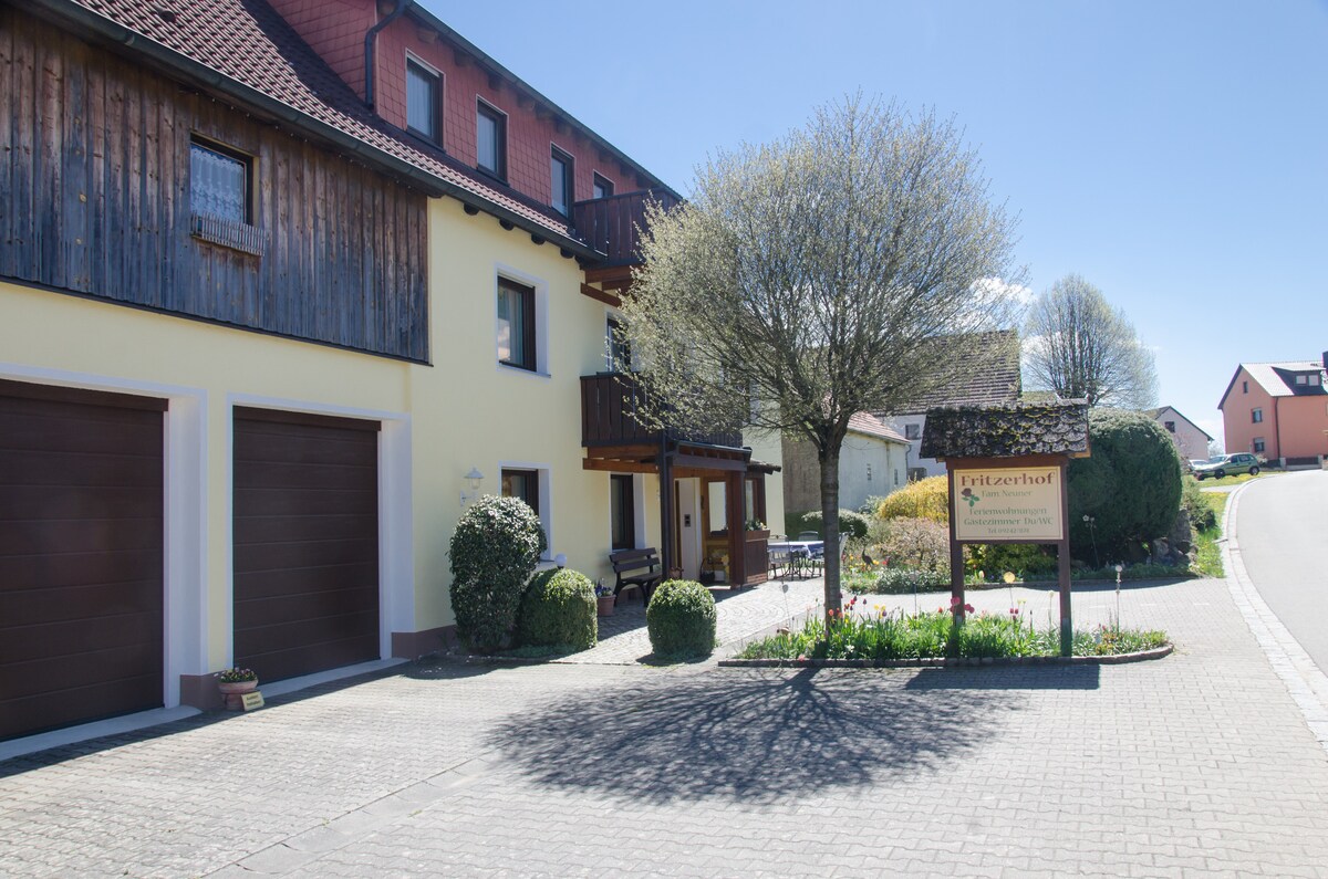 The exterior of Fritzerhof is shown, featuring a two-story building with a combination of wooden and yellow facades. A paved driveway leads to the property, complemented by neatly trimmed shrubs and a sign displaying the name of the property. Trees provide shade in the yard.