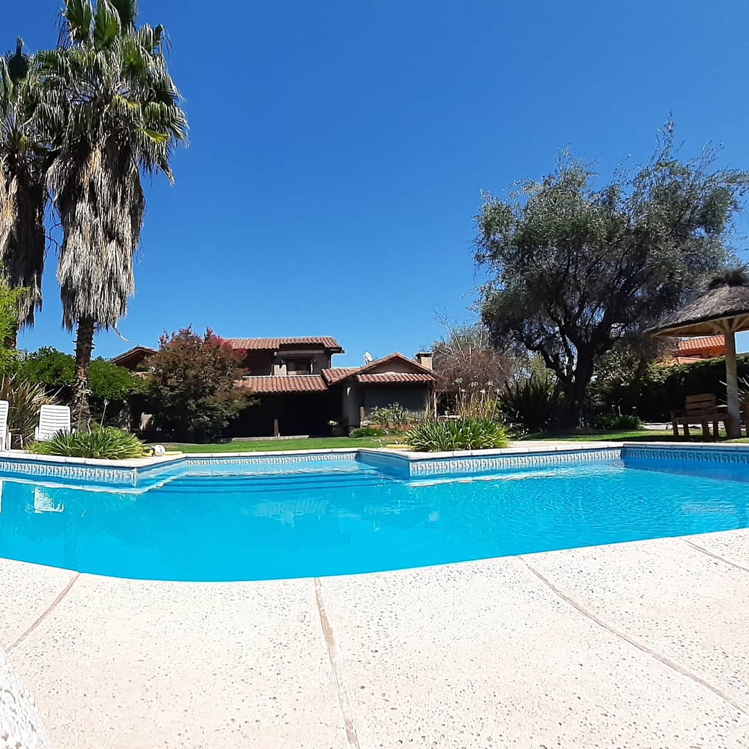 A clear blue swimming pool reflects the bright sky, surrounded by lush greenery and palm trees. In the background, the charming architecture of the main house and outdoor seating area can be seen, providing a serene setting for relaxation.
