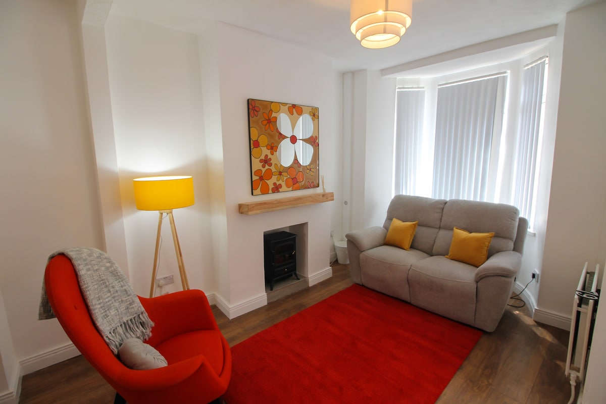A living area features a modern gray sofa with yellow accent cushions and an orange armchair, complemented by a bright red rug. A framed piece of wall art enhances the decor, while a floor lamp provides illumination beside the sofa. Light streams through large windows with vertical blinds.