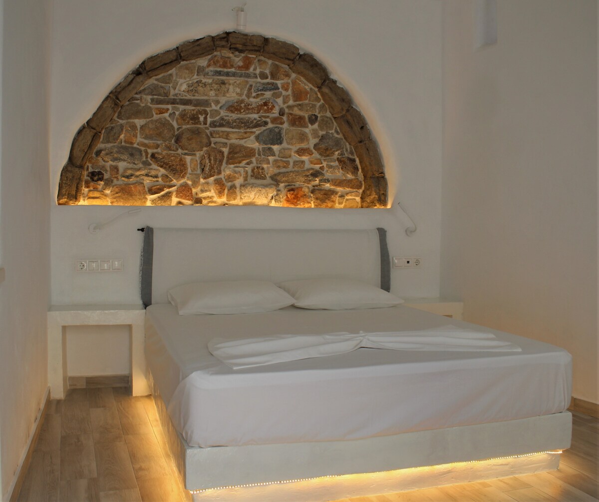 A spacious sleeping area features a comfortable bed with soft linens. An arched stone wall adds unique character, enhanced by subtle lighting underneath the bed. Side tables are placed on either side, providing convenient space for personal items.