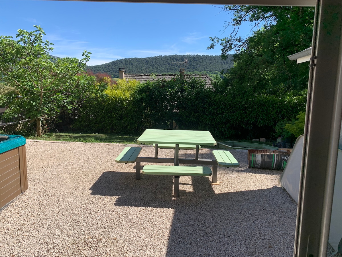 An outdoor space is visible featuring a green picnic table with attached benches, situated on a gravel surface. Surrounding greenery includes trees and bushes, while distant hills are seen under a clear sky.