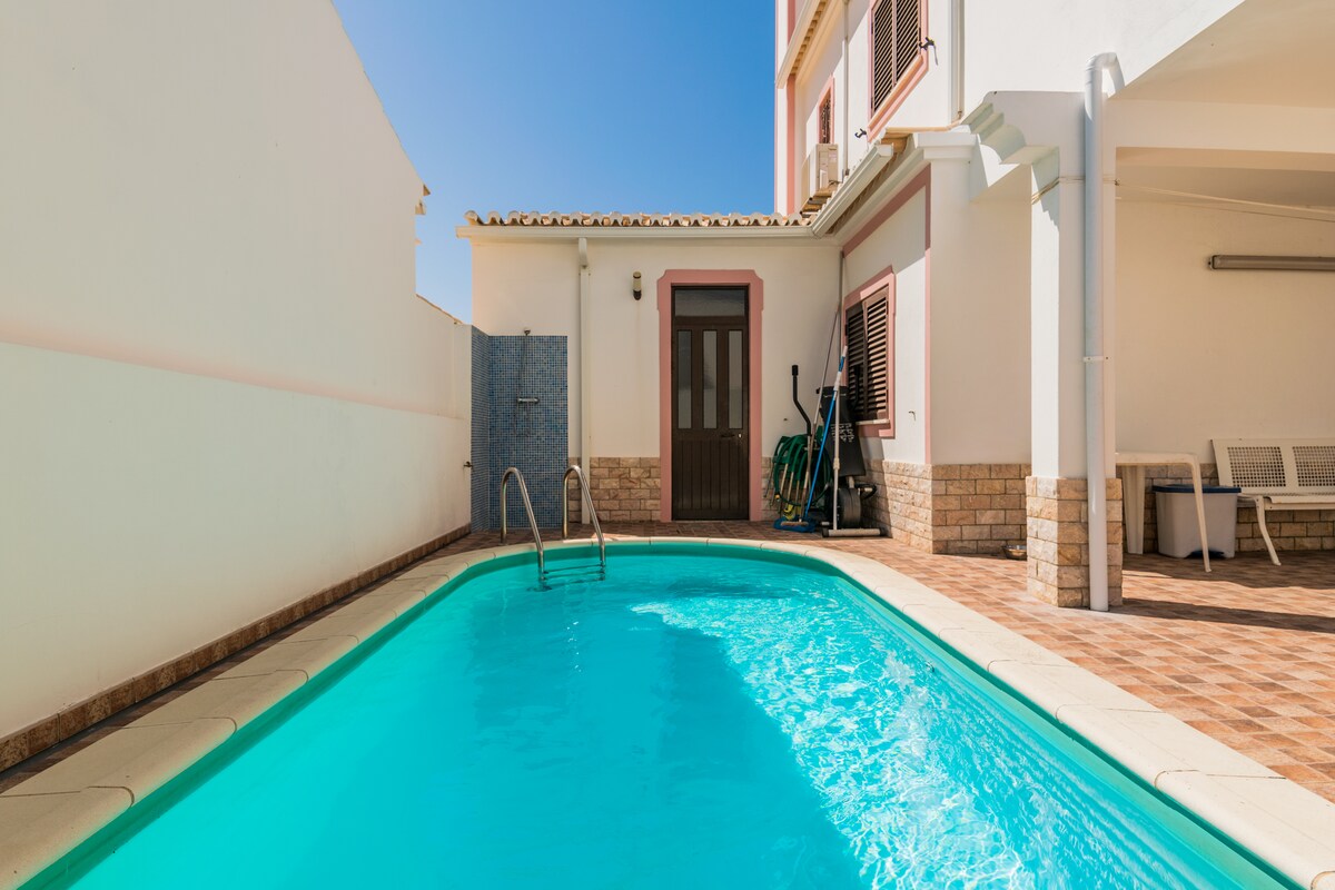 A private swimming pool is framed by tiled decking, featuring steps leading into the water. The area is well-lit, showcasing a clear blue sky above. A door from the pool area leads to the interior, while outdoor seating options are visible nearby.