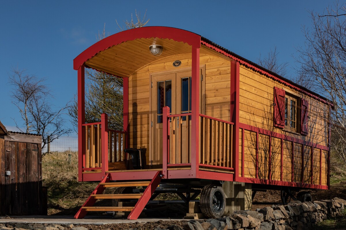 A hand-built gypsy caravan reflects traditional French 'Roulotte' design. The exterior features wooden siding painted in rich hues of red and golden yellow. A spacious front porch with steps invites relaxation, showcasing two upright chairs and welcoming windows adorned with shutters.