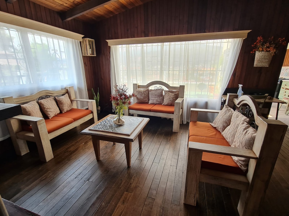 A cozy seating area features wooden furniture with orange accent cushions, positioned around a low coffee table. Natural light filters through sheer white curtains, illuminating the warm wooden floors and adding to the inviting atmosphere.
