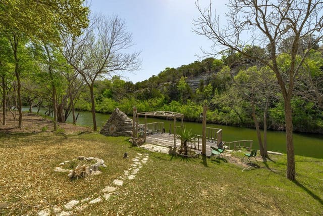 A peaceful riverside area is showcased, featuring a wooden deck with seating arrangements. Lush greenery lines the riverbank, while a stone pathway leads through the grassy space, inviting relaxation amidst nature.