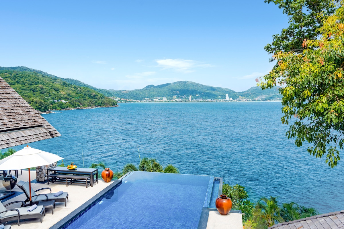 A serene infinity pool offers stunning views of the water and lush hills beyond. Outdoor loungers are arranged around the pool area, complemented by a shaded dining space under a large umbrella. Soft waves are visible in the distance, creating a tranquil atmosphere.
