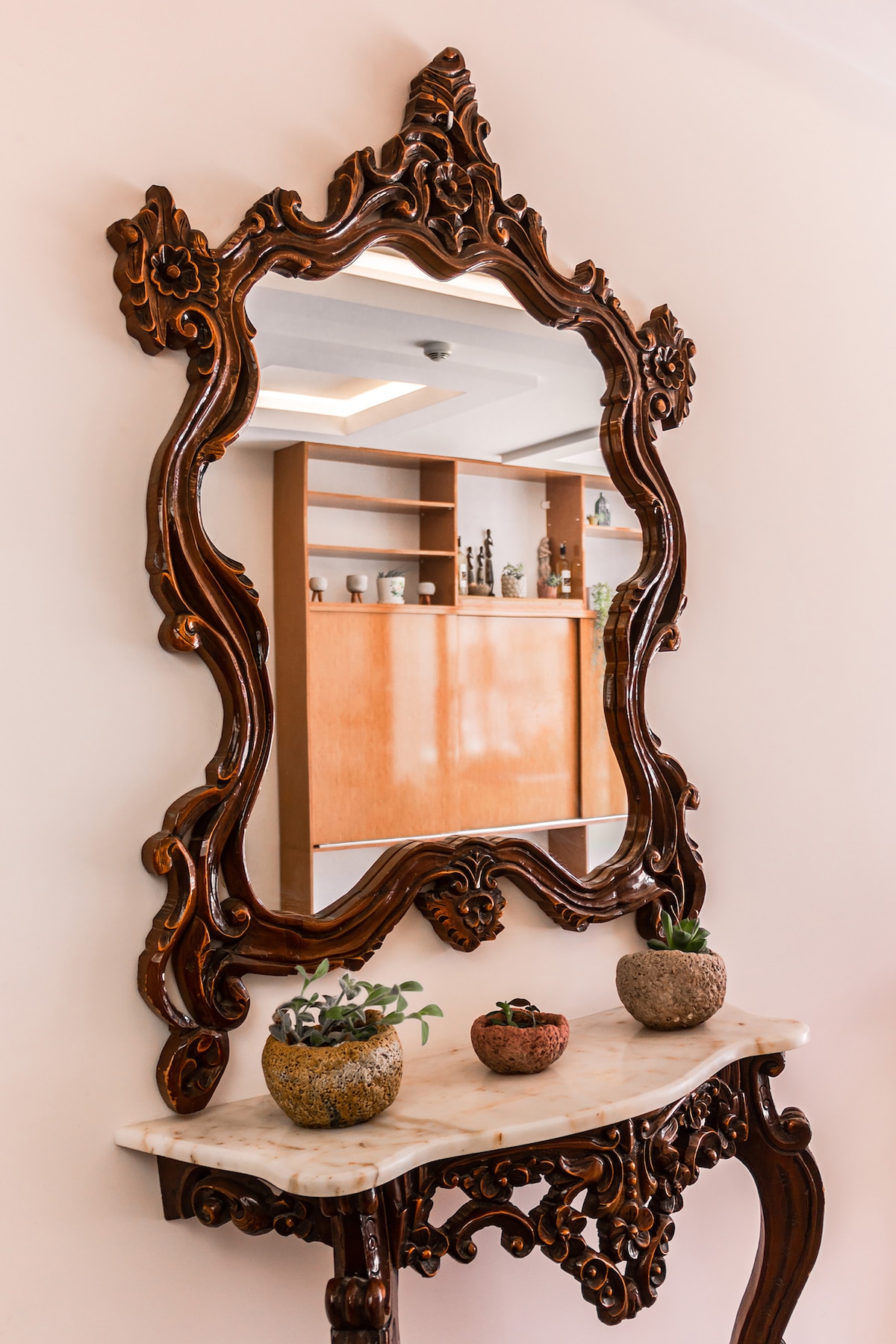 An ornately designed wooden mirror frame is mounted on a light-colored wall, reflecting the surroundings. Below, a marble-topped console table showcases three decorative pots with small greenery, adding a subtle natural touch to the space.