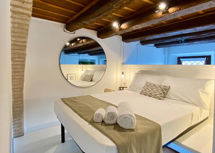 Cama en el loft que parece casi suspendida gracias al apoyo en parte en un piso elevado con respecto al piso. Ambiente cómodo ablandado por resina blanca en contraste con las vigas de madera antiguas.