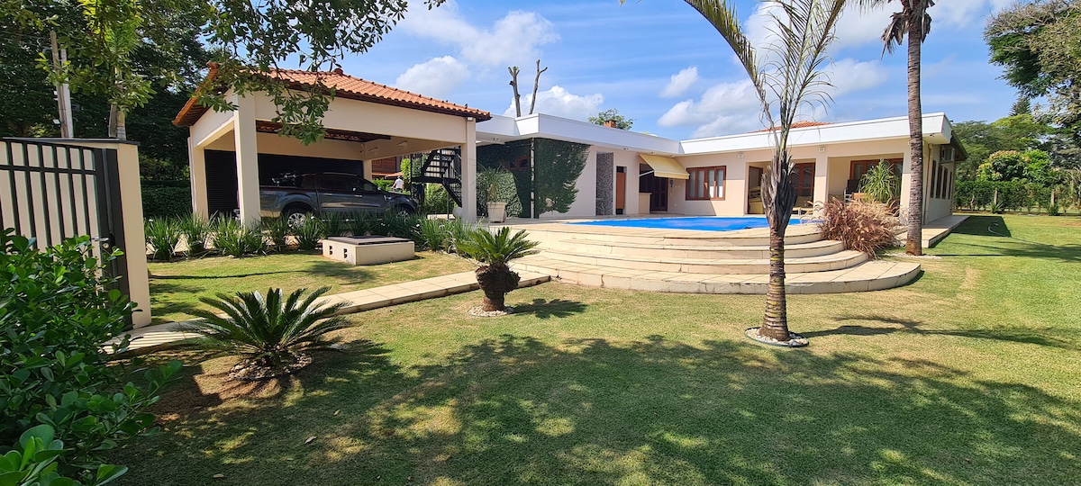 The exterior features a modern house set amidst lush landscaping, with a swimming pool visible in the foreground. Stone steps lead to the entrance, framed by palm trees and greenery. A covered carport offers convenience, contributing to a serene outdoor environment.