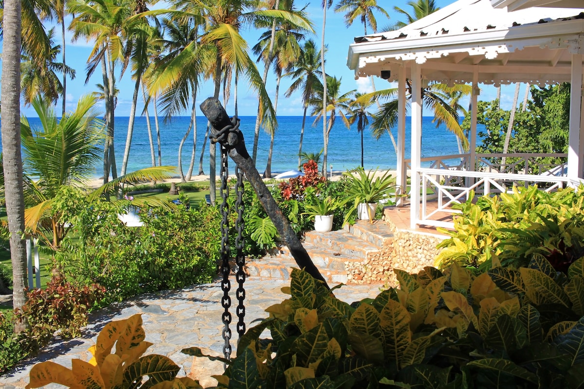 A lush garden setting is visible, framed by vibrant greenery and palm trees. An anchor is prominently displayed in the foreground, leading to a traditional gazebo with a white roof and spacious seating area. The ocean and blue sky can be seen in the background.