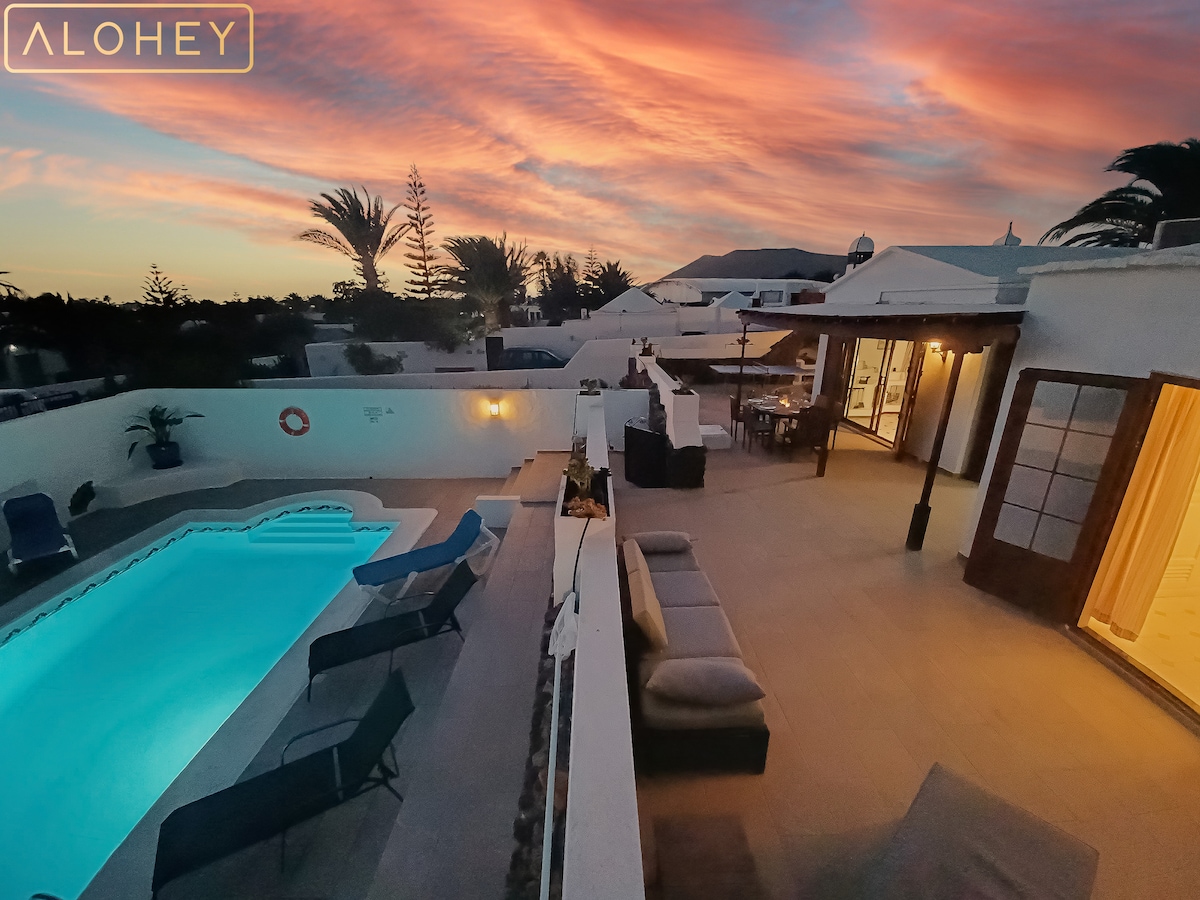 An aerial view reveals a swimming pool surrounded by lounge chairs, offering a space for relaxation. The patio is complemented by comfortable seating and palm trees, with a vibrant sunset sky providing a picturesque backdrop.