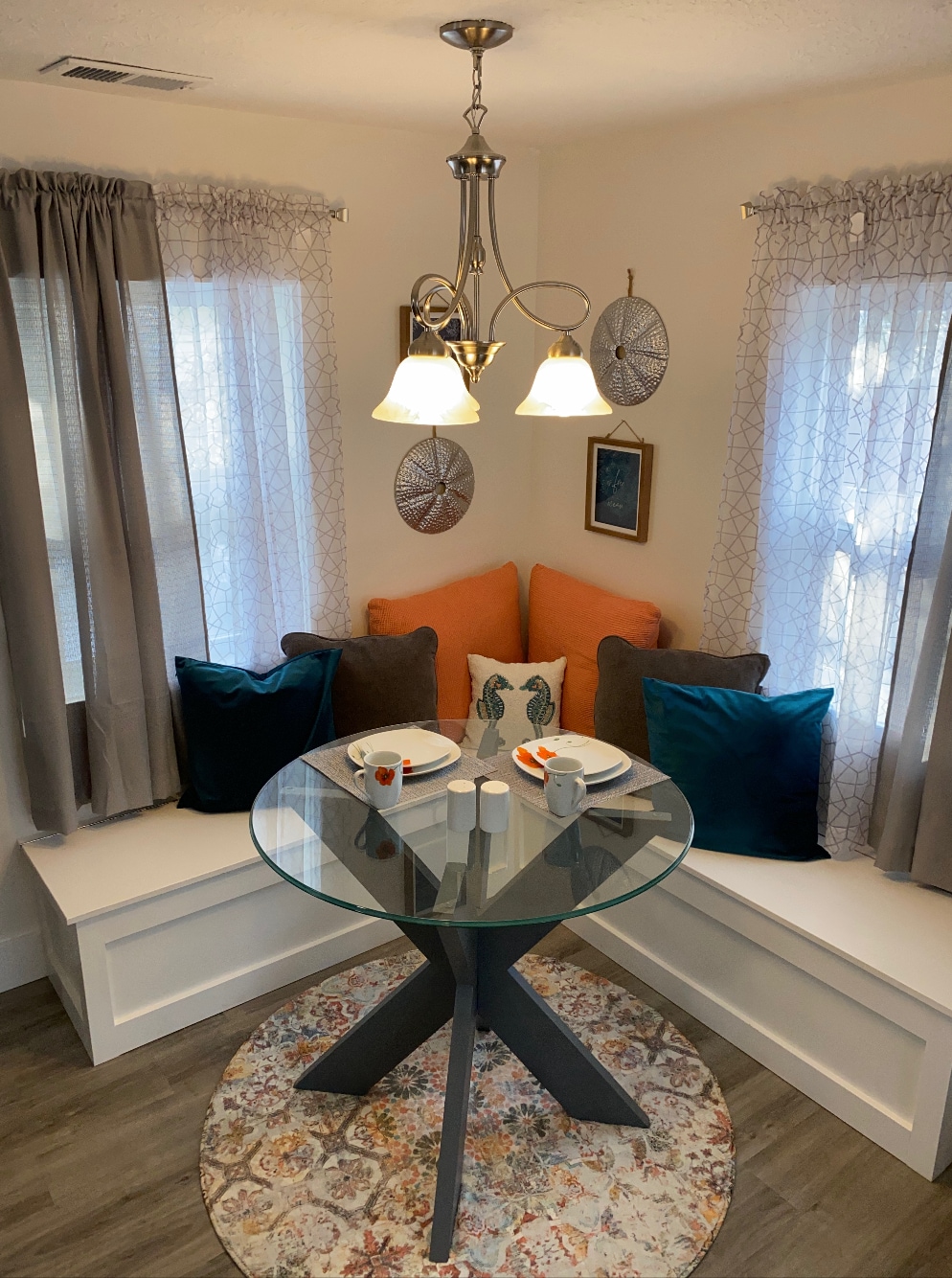 A cozy dining area features a round glass table with two place settings, complemented by colorful cushions on a built-in seat. Soft natural light filters through two windows adorned with sheer curtains, enhancing the inviting space.