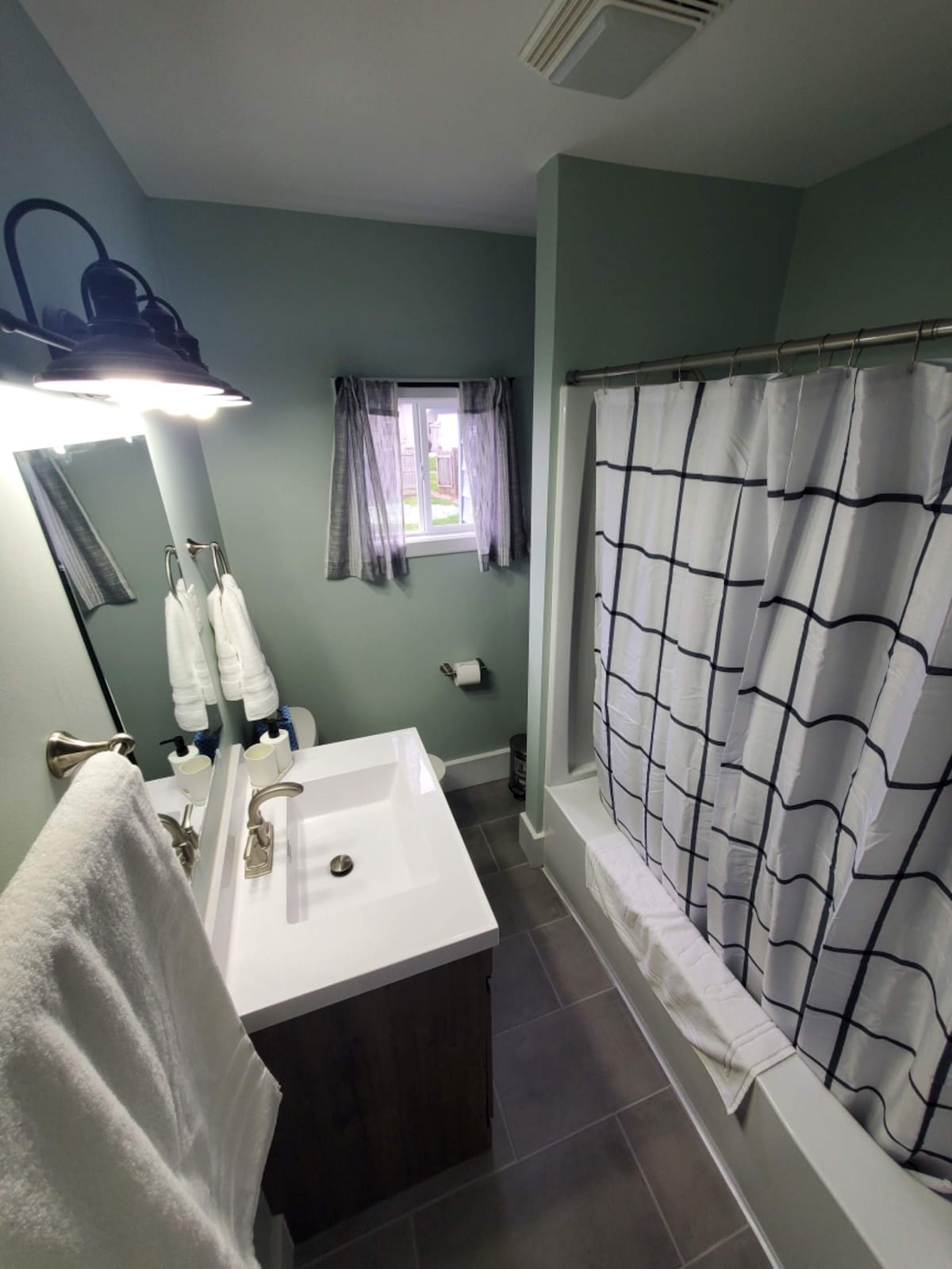 A modern bathroom is depicted, featuring a clean, white sink and a mirror above it. A light fixture illuminates the space, while neatly arranged towels hang on a rack. A shower curtain with a grid pattern encloses the bathtub, and a small window allows natural light in.