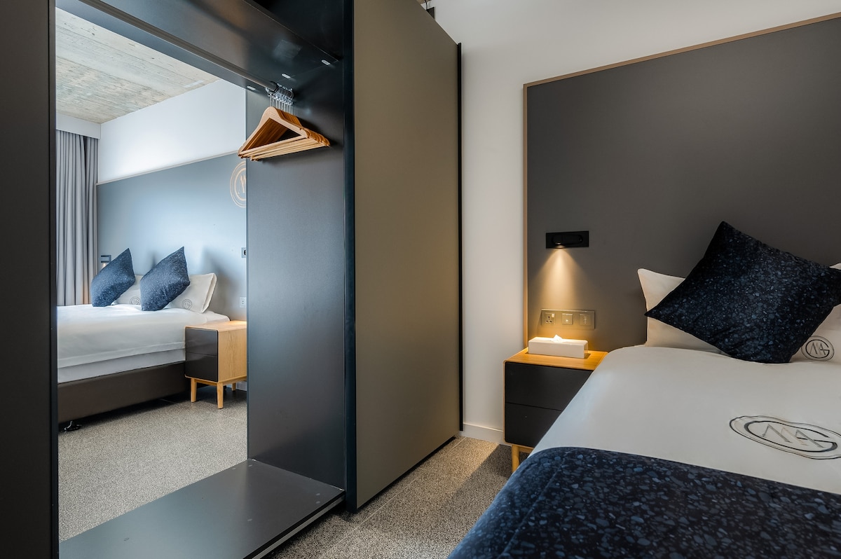 An interior view of the Family Suite showcases two sleeping areas separated by a modern dividing wall. One side features a spacious bed with dark-colored bedding and decorative pillows. A nightstand with a lamp is positioned beside the bed, while the wardrobe is visible nearby.