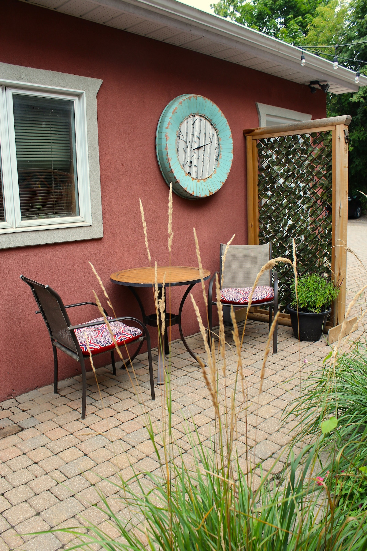 A cozy outdoor seating area features a round table surrounded by two chairs with patterned cushions. A decorative wooden divider adds privacy, and a potted plant is positioned nearby. Tall grasses frame the scene, enhancing the natural atmosphere.