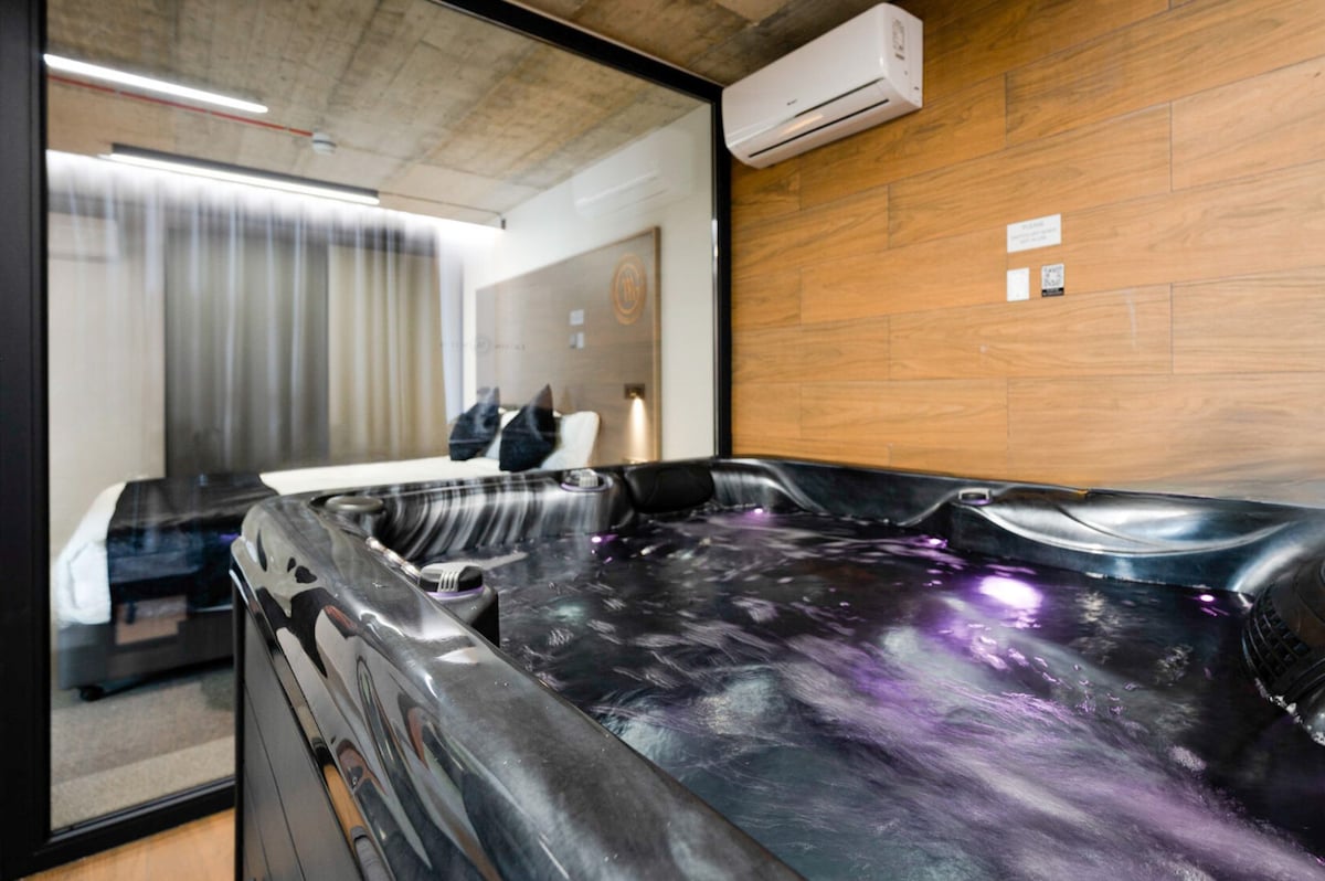 A hot tub filled with bubbling water is featured prominently, with soft purple lighting accentuating its contours. A glass wall separates the tub from a cozy sleeping area, where a spacious bed with dark linens is visible in the background, further enhancing the suite's tranquil ambiance.