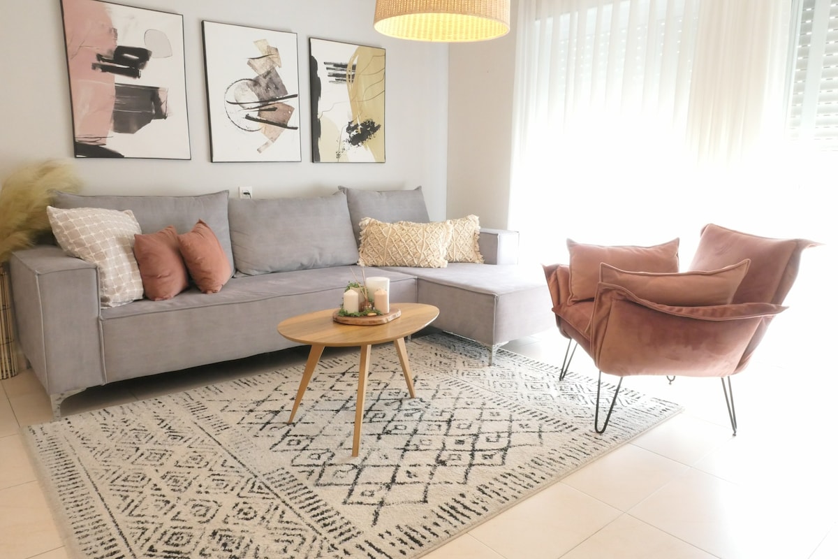 A modern living area is presented, featuring a light gray sectional sofa adorned with decorative cushions. A round wooden coffee table sits on a patterned area rug, and an accent chair in soft pink is positioned nearby. Large windows allow natural light to fill the space.