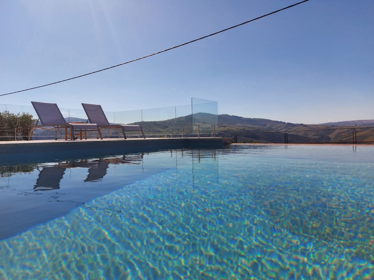 A tranquil infinity pool reflects the clear blue sky, with modern loungers positioned nearby. Glass fencing provides unobstructed views of the surrounding hillside, enhancing the serene environment.