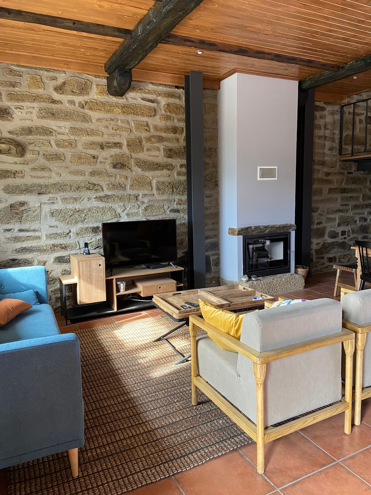 The living area features stone walls and wooden beams, complemented by a flat-screen television and a cozy fireplace. A mix of comfortable seating, including a blue sofa and light-colored chairs, creates a welcoming space. Natural light fills the room through the large windows.