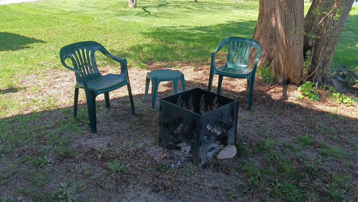 A fire pit surrounded by three green plastic chairs is situated in a grassy area. The fire pit, made of metal, shows remnants of previous fires. A small chair is positioned close by, allowing for a comfortable gathering space.