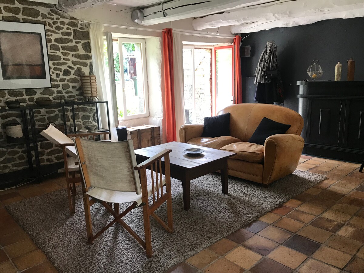 A cozy living area is set with a plush couch and wooden chairs around a simple table. Natural light enters through multiple windows, complemented by soft curtains. Textured stone walls add character, while a neutral rug grounds the space.