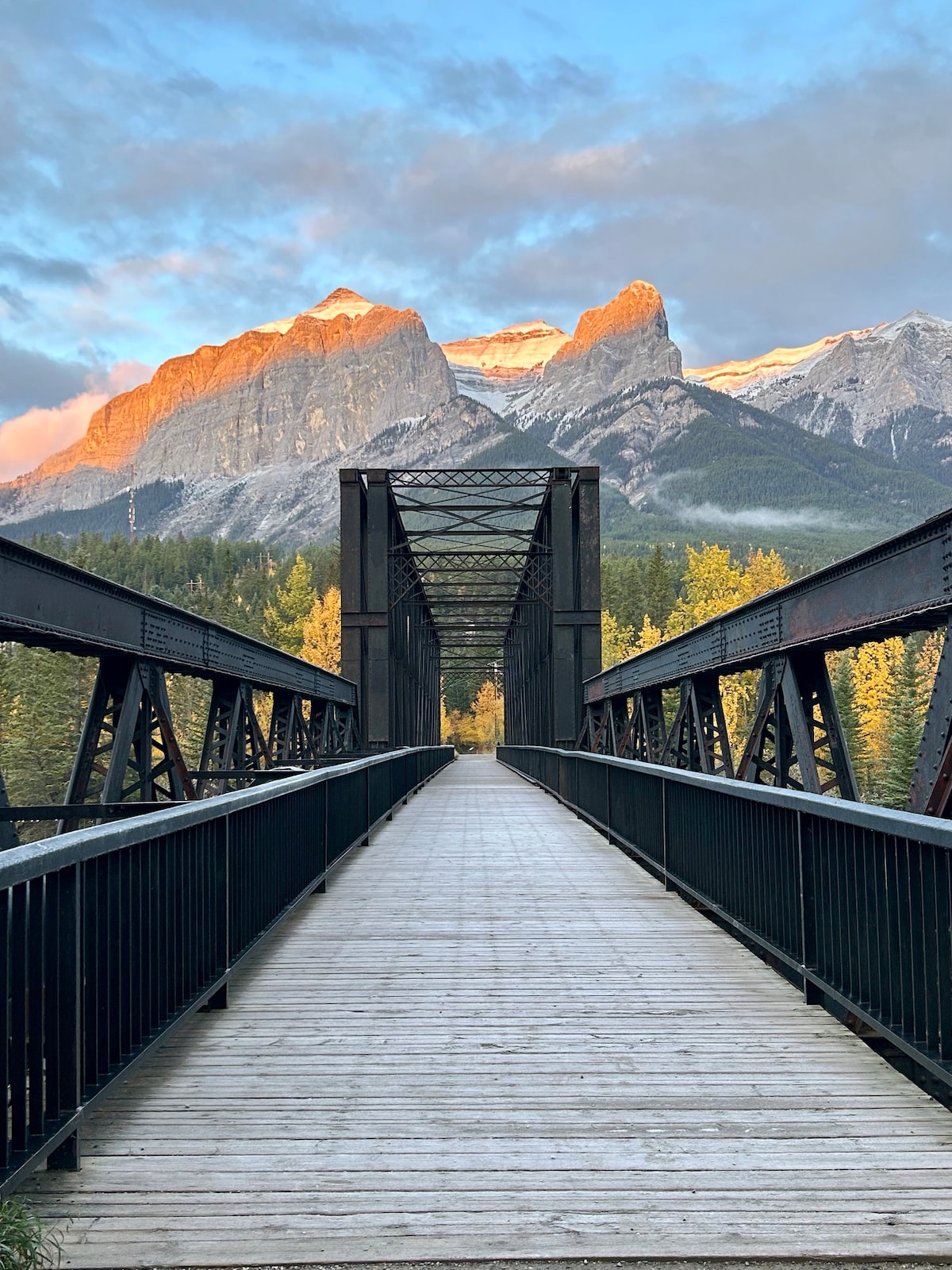 A wooden pedestrian bridge spans a tranquil setting, flanked by rustic black railings. Majestic mountains are visible in the background, bathed in warm early morning light. Lush trees are prominent along the sides, enhancing the natural beauty of the landscape.