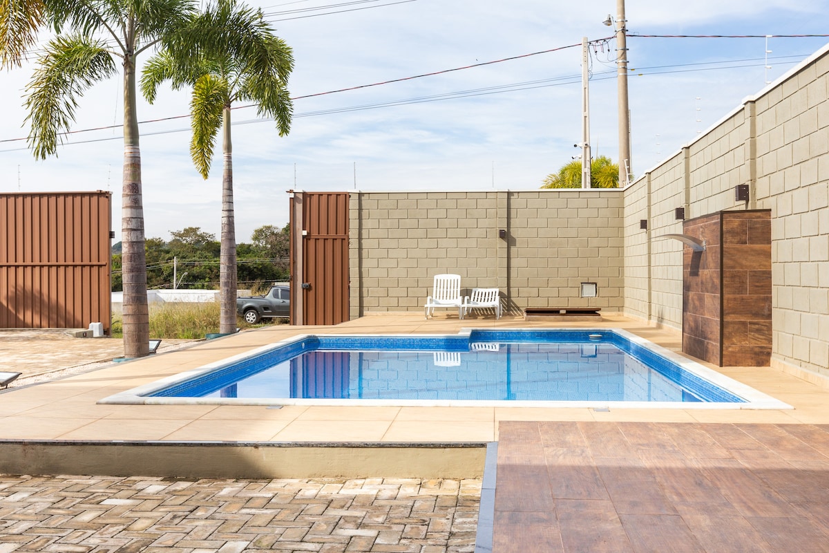 A rectangular pool is framed by a smooth deck and bordered by palm trees. Lounge chairs are positioned nearby, providing a space for relaxation. The surrounding areas feature a clean, modern fence and a mix of paved surfaces, enhancing the overall outdoor ambiance.