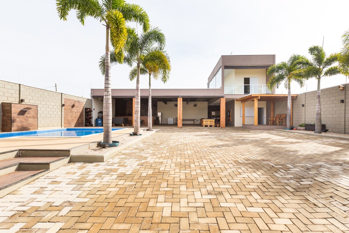 The outdoor area features a spacious paved patio bordered by palm trees. A swimming pool is visible on the left, while a two-story building is positioned in the background. A shaded terrace area is included, providing seating options for relaxation.