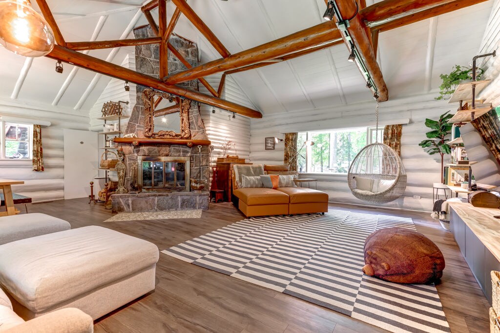 A spacious living area features a high-beamed ceiling and a large stone fireplace as a focal point. A comfortable seating arrangement includes a sofa and a hanging chair. Natural light filters through windows, highlighting the decor and the unique striped rug below.
