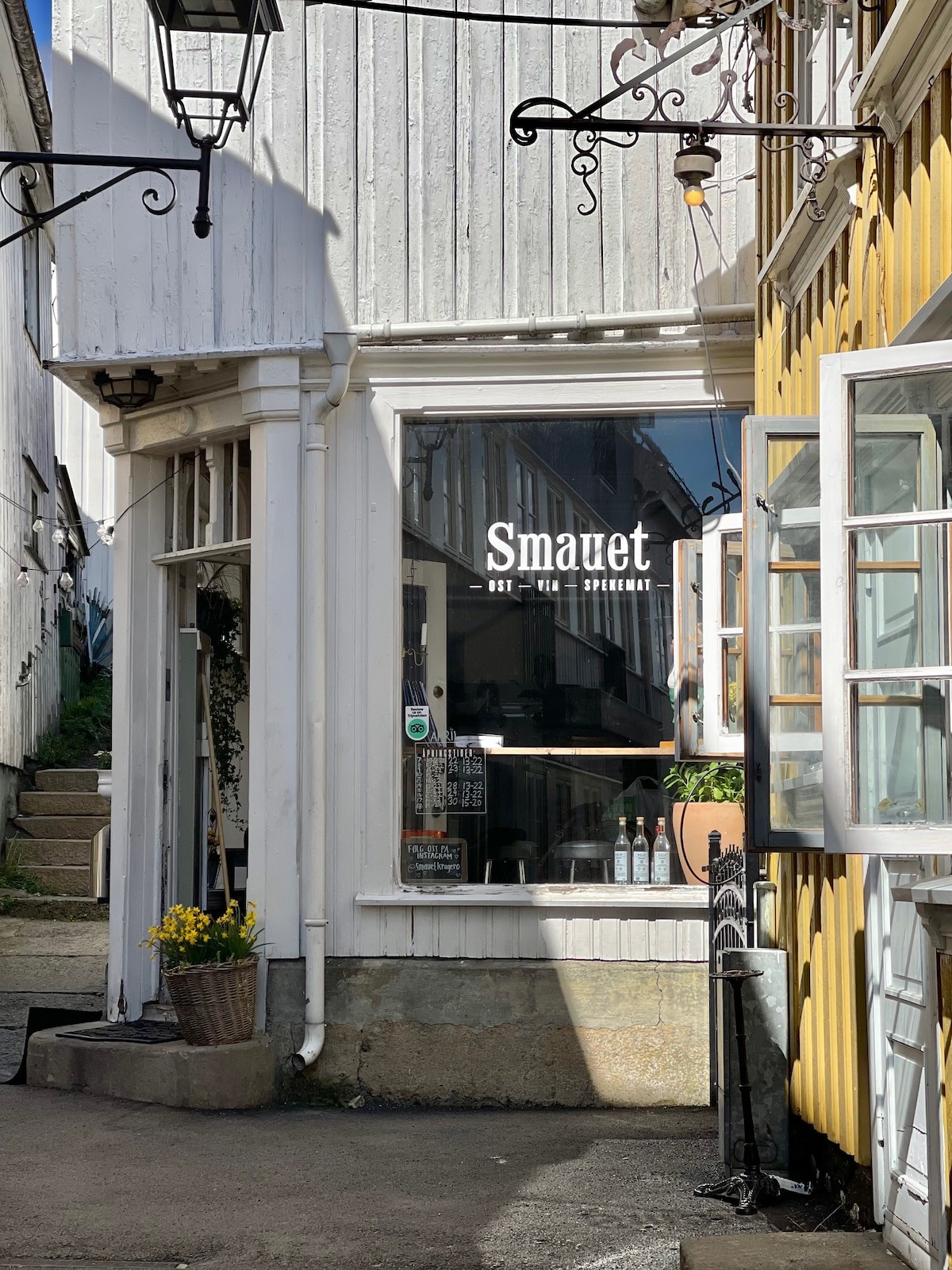 The image showcases the entrance of a quaint eatery with large windows allowing natural light to fill the space. A sign indicates the name 'Smauet.' A flower pot sits outside, complementing the surrounding charming architecture of the building.