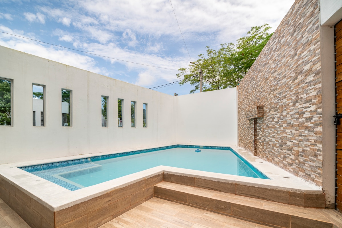 A private pool area is depicted, featuring a crystal-clear water surface surrounded by smooth wooden decking. Light-colored walls reflect natural light, with tall windows allowing views of the greenery outside. The pool is designed with steps leading into the water for easy access.