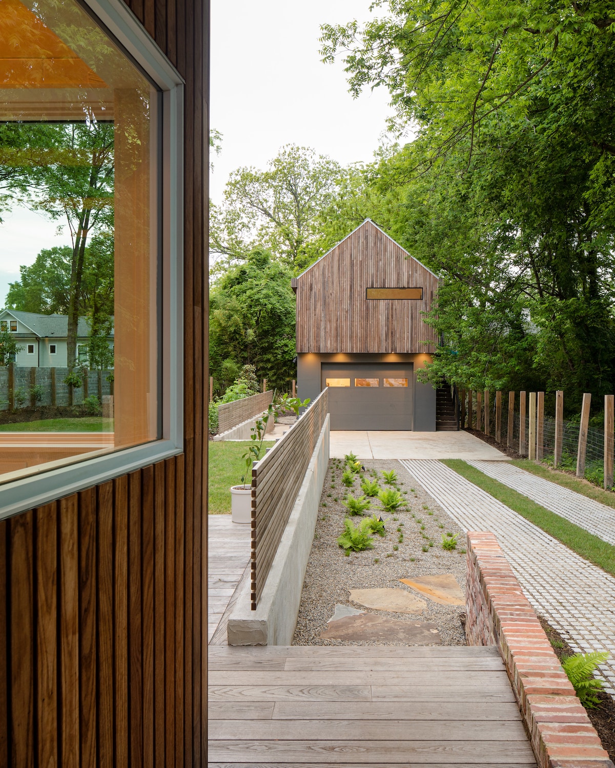 A modern, detached garage is visible at the end of a paved driveway, framed by greenery. The garage features a wooden façade and sleek design, complemented by a well-maintained garden area with gravel pathways and low plants. The inviting entrance is marked by wooden steps leading towards the house.