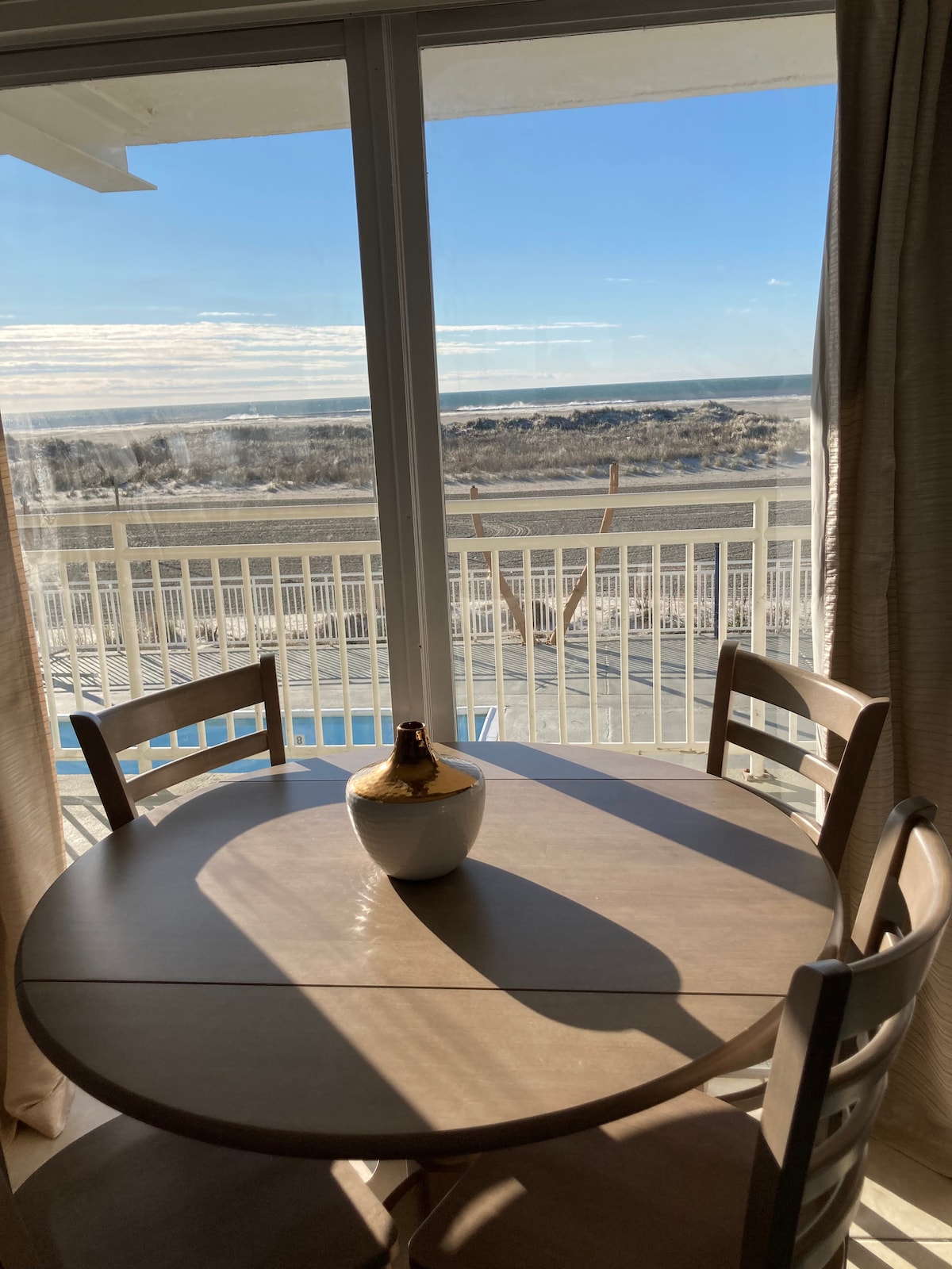 A round wooden dining table with two chairs is positioned near a large window. Bright sunlight streams in, illuminating the space. Outside, a view of the beach and ocean is visible, framed by the glass doors leading to a balcony.