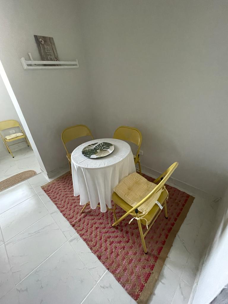 A small dining area is presented with a round table covered by a white tablecloth, accompanied by three yellow chairs. A decorative plate rests on the table, and a textured area rug lays underneath, adding a touch of warmth to the space.