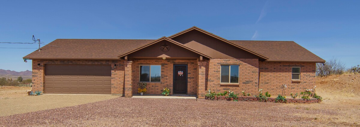 The front view of a single-story brick home is depicted, with a welcoming entrance and large windows on either side of the door. A two-car garage is visible to the left. Desert landscaping surrounds the home, featuring low maintenance plants and a gravel driveway.