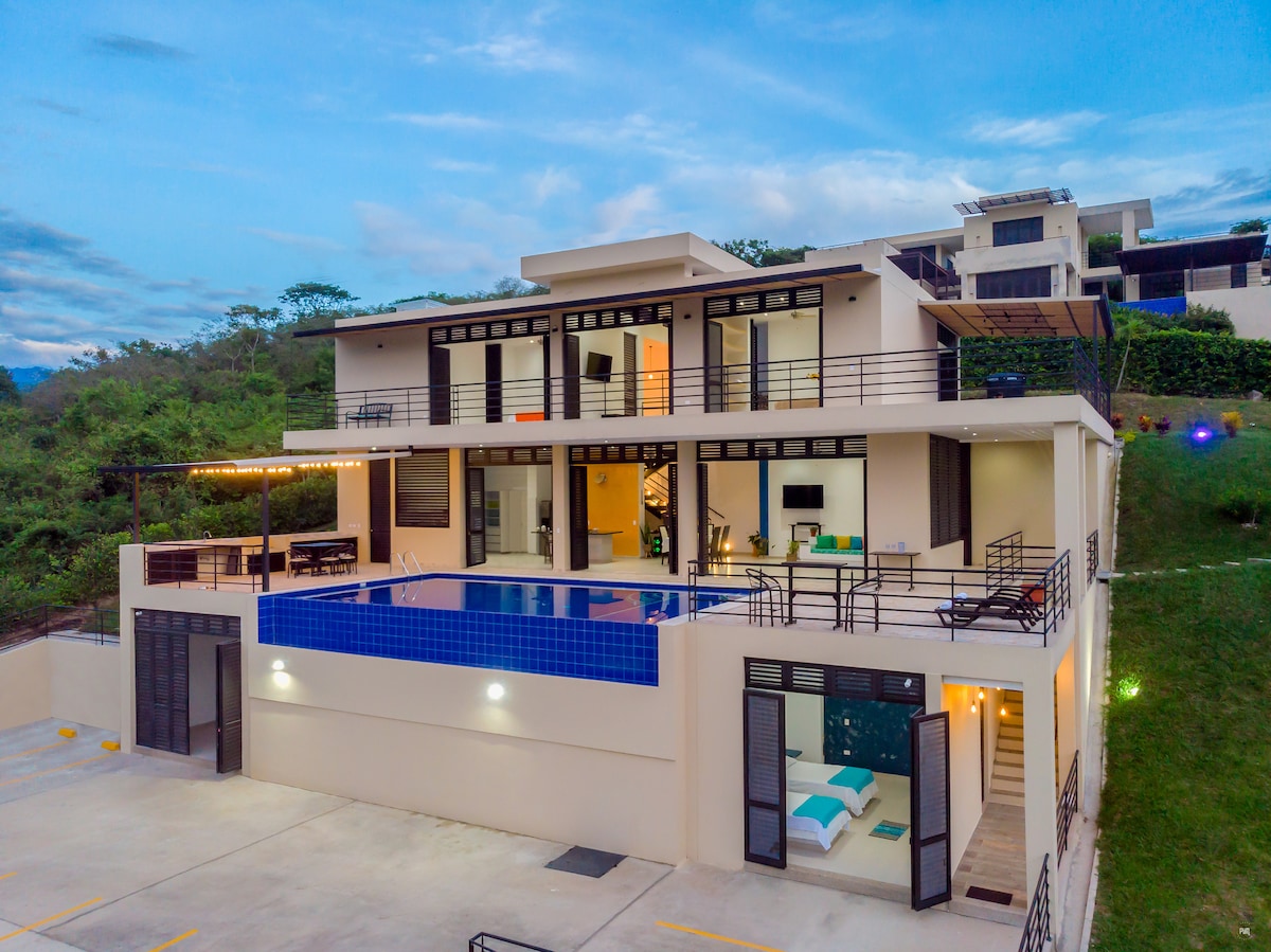 A modern two-story villa is shown, featuring a spacious terrace with an infinity pool. Large glass doors provide visibility into the living areas. The structure is surrounded by green hills and gardens, showcasing a harmonious blend of outdoor and indoor spaces.