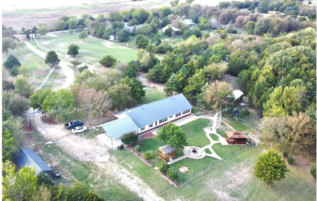 An aerial view showcases a spacious property featuring a main house with a green roof, surrounded by ample greenery and trees. A lawn and small pathways connect various outdoor areas, including a gazebo and play structure. The setting offers a serene countryside atmosphere.