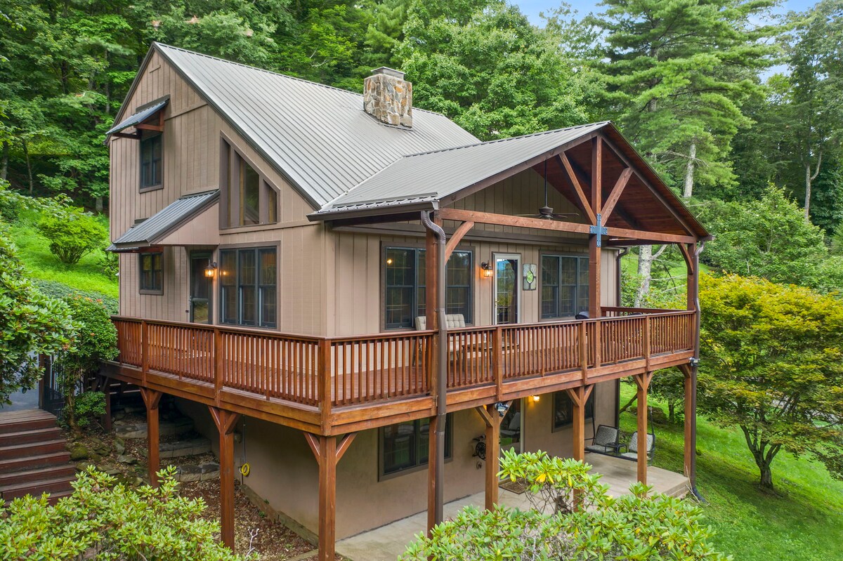 A charming cottage is set among lush greenery, featuring a warm wood deck supported by sturdy posts. The exterior showcases a blend of earthy tones and large windows, while a stone chimney adds a rustic touch. The surrounding trees enhance the serene atmosphere.