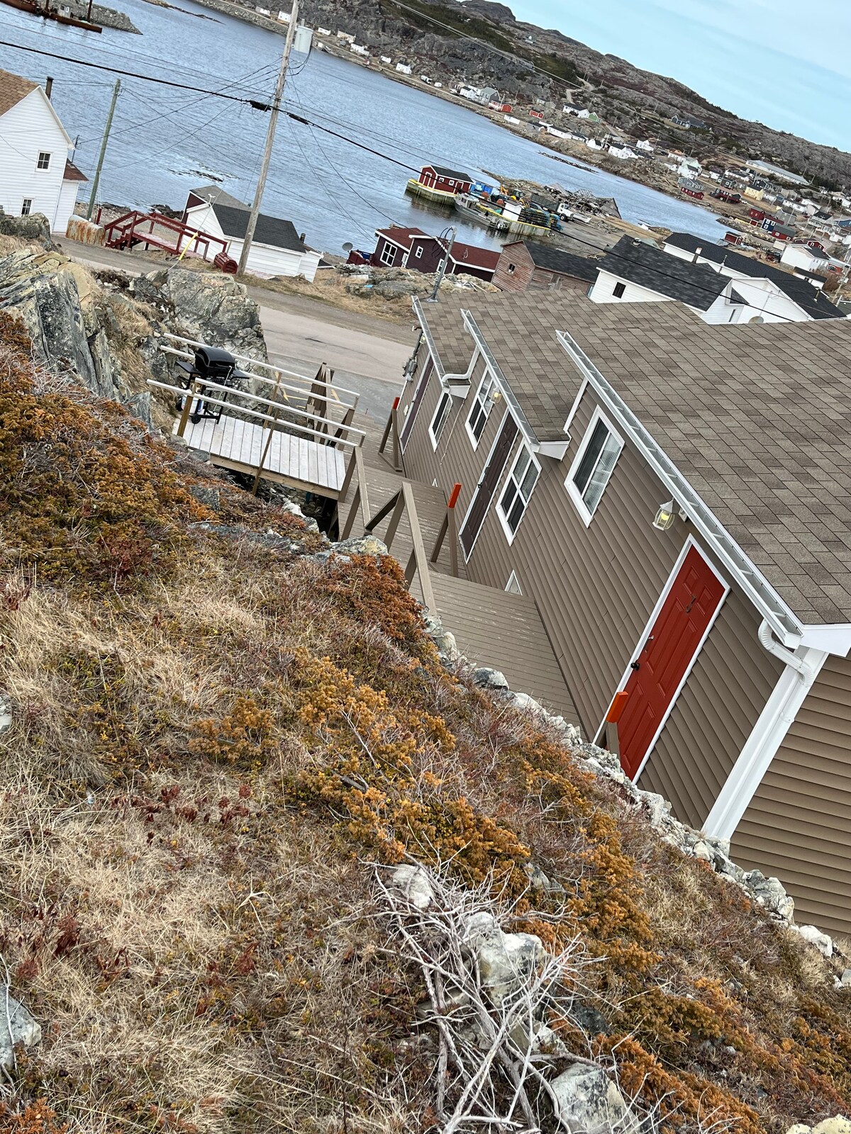 A view from above shows the exterior of a brown-clad building descending a hillside towards Fogo harbour. The nearby landscape is characterized by rocky terrain and low vegetation, with colorful homes visible along the waterfront.