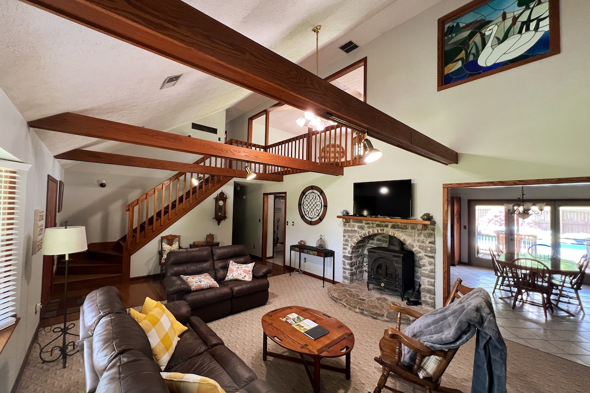 A spacious living area features high wooden beams and a stone fireplace, complementing the comfortable seating arrangements. Natural light floods in through large windows, highlighting the open layout that connects to the dining area and a view of the pool.