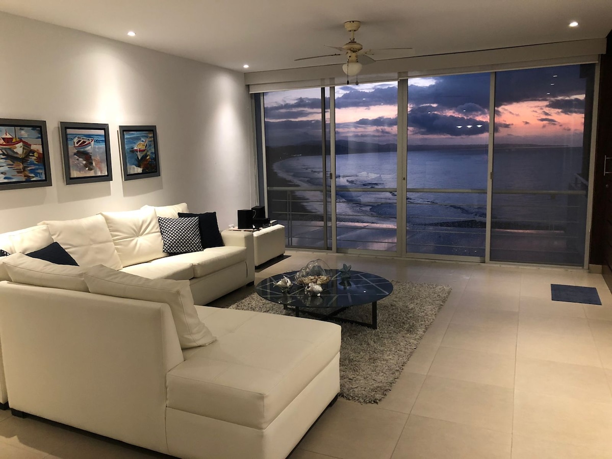 A modern living area is showcased with a white sectional sofa and a glass coffee table centered on a soft area rug. Large windows provide a view of the ocean and a sunset, while framed art pieces adorn the walls.