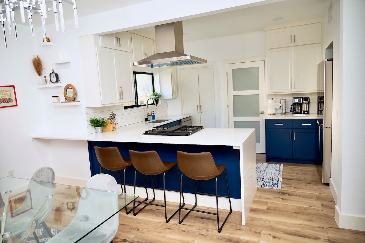 A modern kitchen features a central island with three bar stools and sleek cabinetry in white and navy. Stainless steel appliances and a glass hood highlight the cooking space, while natural light streams in through the window, complementing the warm wooden flooring.