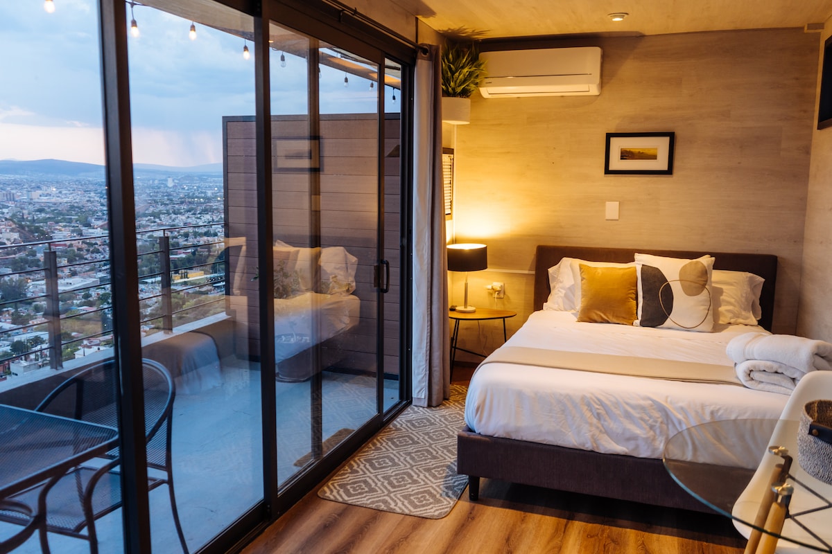 An inviting room features a spacious bed dressed in neutral linens, complemented by decorative pillows. Floor-to-ceiling glass doors open to a private terrace offering panoramic views. A small dining table and chairs occupy one corner, while a lamp provides soft lighting, enhancing the cozy ambiance.