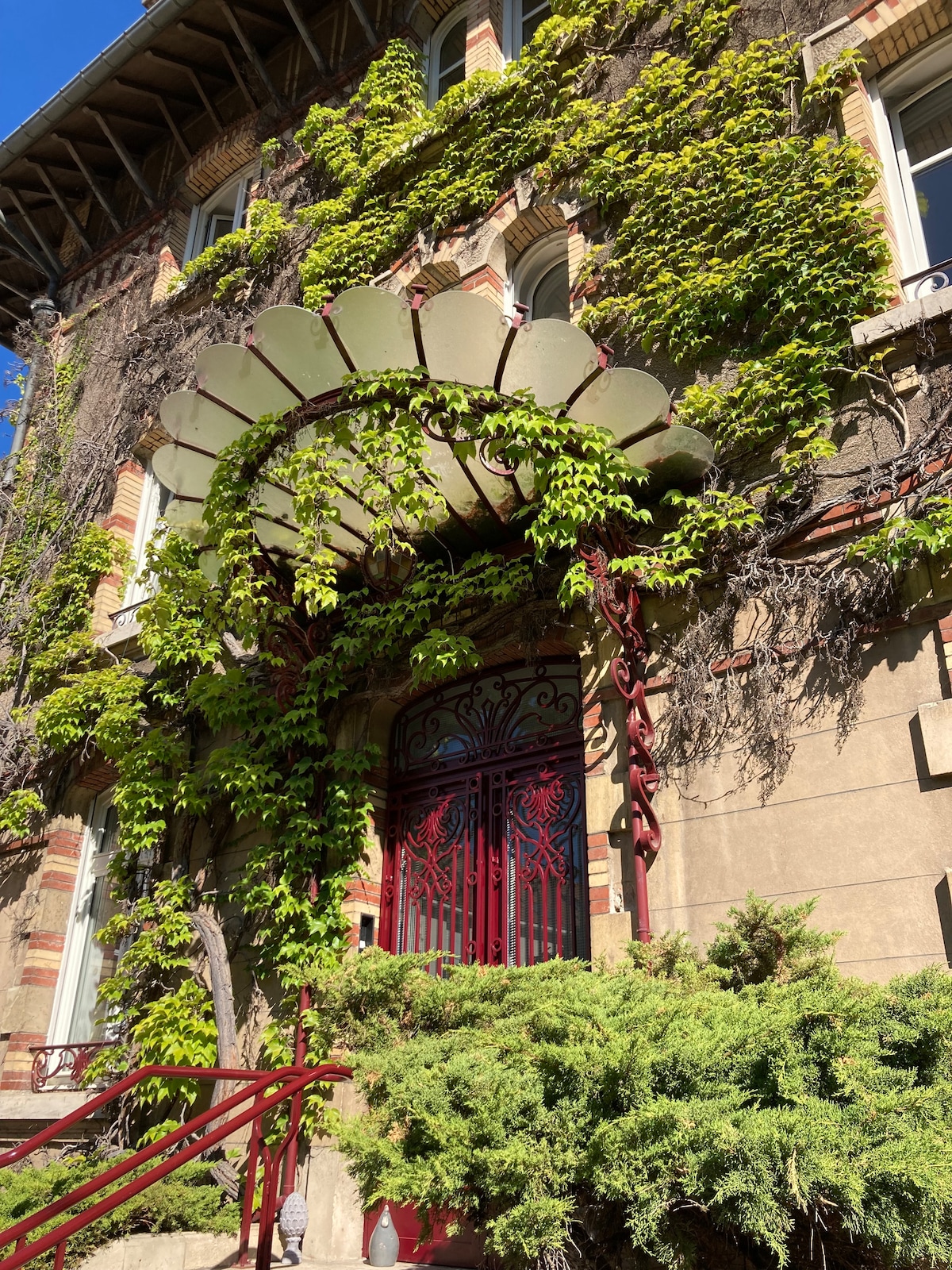 A historical villa is portrayed, featuring intricate ironwork around the entrance, which is adorned with lush green vines. A curved awning extends from the entryway, providing a charming focal point against the stone façade, surrounded by well-maintained greenery.